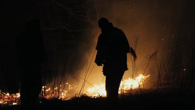 Two firefighters with fire flappers extinguish a fire in the forest at night