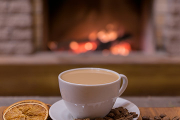 Cup of coffee with milk and coffee beans around the cup, candied fruit before cozy fireplace.