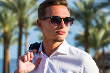 Portrait of a young handsome man in a white shirt and suit stands at the palm trees and sea background.