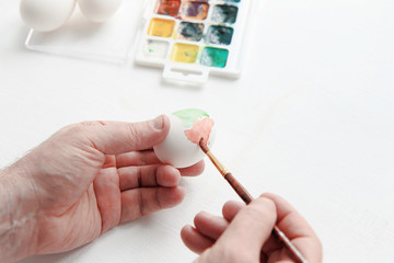 men's hands hold a brush and paint eggs with watercolors. Nearby are watercolor paints and a few eggs on a white background. Easter preparation concept.