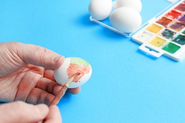men's hands hold a brush and paint eggs with watercolors. Nearby are watercolor paints and a few eggs on a blue background. Easter preparation concept