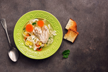 Chicken noodle soup, potatoes, carrots in a green plate served with fried toasts on a dark rustic background. Top view, copy space.
