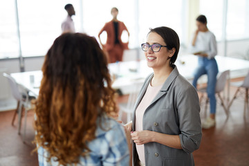Waist up portrait of young female professional smiling happily while talking to colleague in modern...