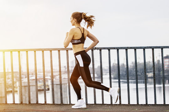 Beautiful fitness girl in stylish sportswear running over bridge with urban view on background. Slender young sports woman jogging in city. Attractive girl in motion. Doing workout, fitness outdoors
