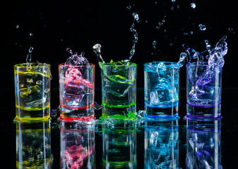 Multicolored glasses filled with alcoholic drinks, with splases of ice cubes falling inside, standing on the mirror surface. Black background. Conceptual, celebrated, commercial design