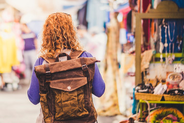 Obraz premium Blonde curly hair woman backpacker traveler viewed from rear at used market enjoying the shopping and alternative vacation - leather backpack view for travel concept
