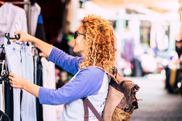 Cheerful traveler young blonde curly woman looking and choosing clothes at the used market during...