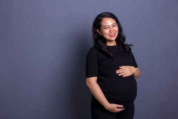 Portrait of happy attractive Asian mother with her pregnant belly smiling at camera, standing...