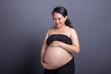 During maternity. Attractive Asian childbearing woman in black clothes caressing her big belly. Over grey background