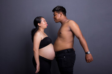 Fun pregnancy concept. Pregnant wife and her husband joking around, their belly bump against each other. Over grey studio background