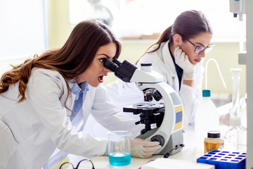 Woman in laboratory looking surprised on microscope with other working woman beside her