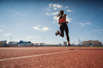 From below back view of teen girl in sportswear sprinting on running track at stadium under blue...