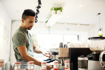 Side view of young concentrated bartender pouring coffee into cup from espresso machine at counter in small coffee shop - obrazy, fototapety, plakaty