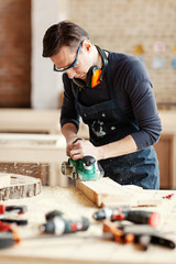 Young woodworker sanding wooden plank with flatbed sander machine, his workbench covered with tools, wood shavings and sawdust