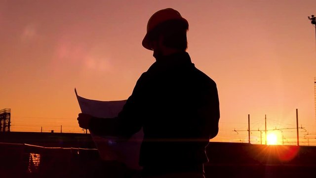 Sagoma di spalle di un uomo architetto con caschetto di protezione in testa visto di spalle al tramonto. Osserva il progetto sul foglio e analizza se il lavoro in cantiere si sta svolgendo bene.