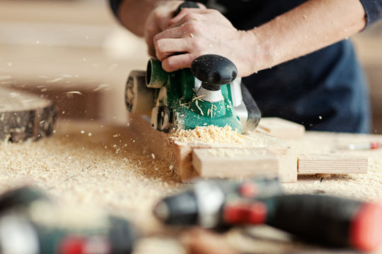 Closeup crop hands of young woodworker making wooden detail and using electric sanding machine at workplace covered with sawdust near equipments