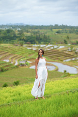 Fototapeta na wymiar Tanned beautiful woman in white long dress smiling and posing against the background of rice fields