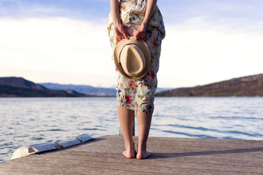 Back view of barefoot young woman standing on jetty holding summer hat, partial view