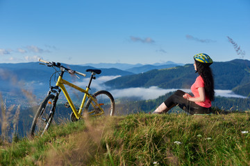 Happy attractive woman biker sitting on the top of mountain near yellow bicycle in the morning, wearing helmet and red red t-shirt. Foggy mountains, forests on the blurred background. Copy space