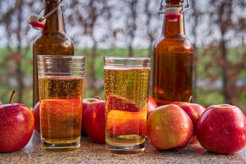 Close up Picture on two glasses or jars full of organic sparkling apple cider and two rustic bottles with red riped apples on stone garnit table in the garden restaurant during summer sunny evening.