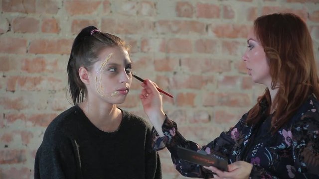 Close-up woman professional makeup artist makes wax plastic makeup for art cinema and paints face of young cute girl and creates image for shooting scene on background brick wall