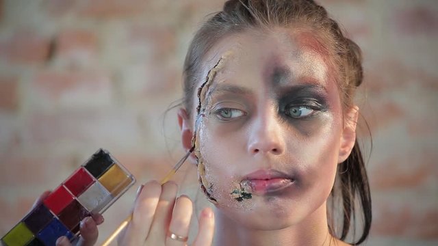 Close-up woman professional makeup artist makes wax plastic makeup in form of bloody wound for art cinema and paints face of cute girl and creates image for shooting scene on background brick wall