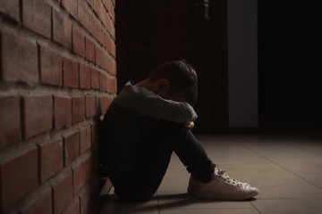 Sad little boy sitting on floor near brick wall indoors