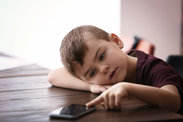 Naklejka premium Sad little boy with mobile phone sitting at table indoors