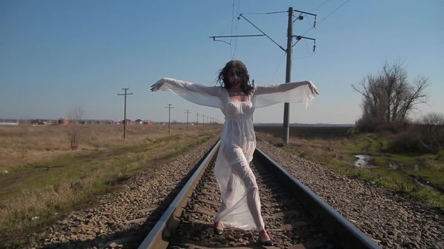 Camera motion, amazing slim unusual girl in white translucent dress and with art plastic wax makeup on her face in form of torn bloody wound makes unusual dance moves on railway tracks in sunny day