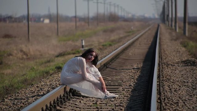 Amazing slim unusual beautiful girl in white translucent dress blown by wind and with wax makeup on face in form of bloody wound sits on railroad tracks in waiting