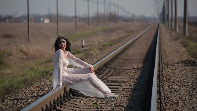 Amazing slim unusual beautiful girl in white translucent dress blown by wind and with wax makeup on face in form of bloody wound sits on railroad tracks in waiting