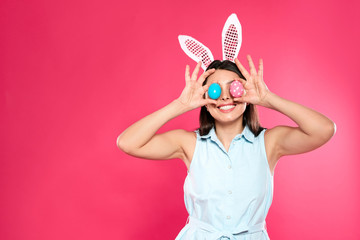 Beautiful woman in bunny ears headband holding Easter eggs near eyes on color background, space for text