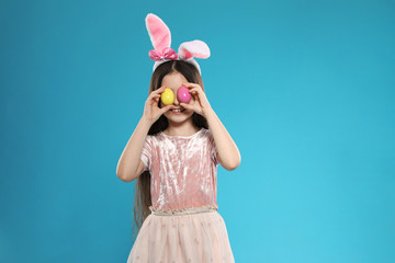 Obraz na płótnie Canvas Little girl in bunny ears headband holding Easter eggs near eyes on color background