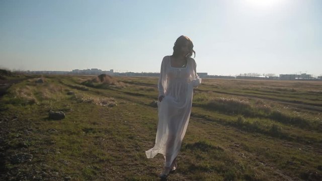 Camera motion, amazing slim unusual girl in white translucent dress and with wax makeup on face in form of bloody wound walks in field and makes beautiful unusual dance moves under boundless blue sky