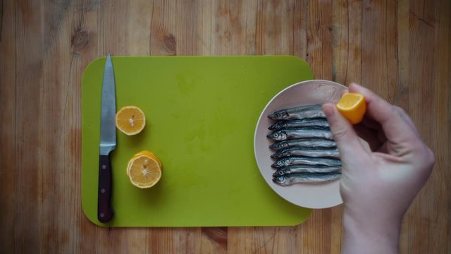 Top view of the process of making a small silver fish, male hands poured fresh lemon juice to capelin, lying in a beige plate on a wooden table.