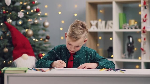 Serious concentrated little boy with a tie on his neck actively drawing with colored pencils shining Christmas tree on the background. Holidays, positive emotions, happy childhood, art