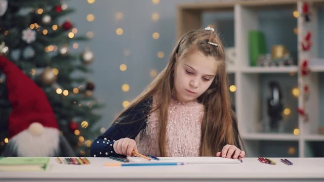 Close-up of lovely cute little girl draws the pictures using color pencils in the album in the art class on Christmas tree background. Coziness, positive mood, presents, holidays. Slow motion