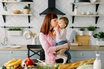 Cute little baby girl eating a carrot in the arms of her mother in their kitchen. Happy mother cooking with her baby girl in light kitchen. Family, food, eating, breakfast and people concept