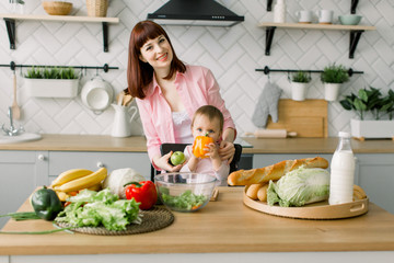 Happy mother with little baby girl in the kitchen. Beautiful young mother holding green apple and her cute baby girl with a yellow pepper sitting on the chair at kitchen.