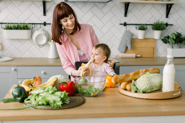 Cute young mother in pink shirt giving a banana to her baby girl sitting on the chair at the table with vegetables and fruits in the kitchen
