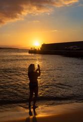 Fototapeta premium Vertical image with a young girl taking pictures with the smartphone on the beach of Cefalu town in Sicily island at sunset