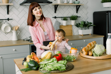 Cute young mother in pink shirt giving a banana to her baby girl sitting on the chair at the table with vegetables and fruits in the kitchen