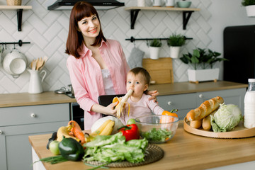 Cute young mother in pink shirt giving a banana to her baby girl sitting on the chair at the table with vegetables and fruits in the kitchen