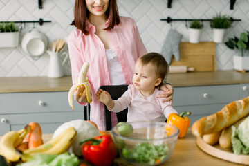 Happy loving family, mother and daugher in the kitchen. Beautiful young mother feeding her cute baby girl with a banana at kitchen. Little baby girl eats banana sitting at the table