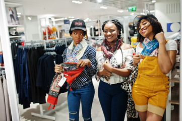 Three african woman choosing clothes at store. Shopping day. They buying caps and scarfs and looking at mirror.