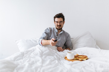 Young man in bed watching tv and having breakfast