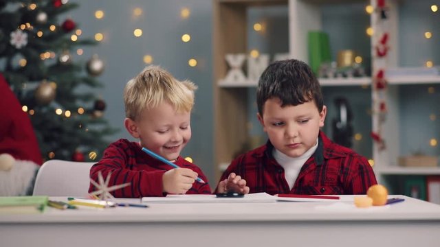 Two cheerful smiling little kids at the table draw with pencils and actively communicate on Christmas tree lights background. Childhood activities, art, creativeness concept. Having fun.