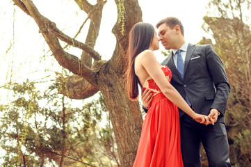 Happy romantic couple are keeping their hands, looking to each other and standing near tree in the park outdoor