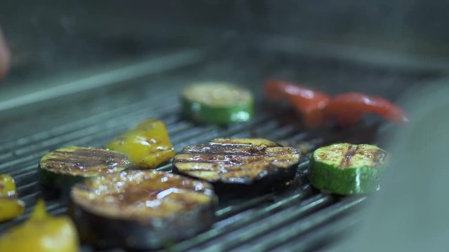 Chef hand turning to the other side vegetables tomato eggplant, zucchini, yellow bell pepper on the grill using metal tongs close up. Cook preparing vegetarian food. Healthy lifestyle