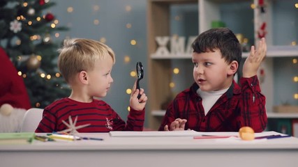 Adorable little blonde boy looking at his talking friend through the magnifying glass. Indoor activities, Christmas tree on the background. Having fun, holidays, true friendship. Games concept